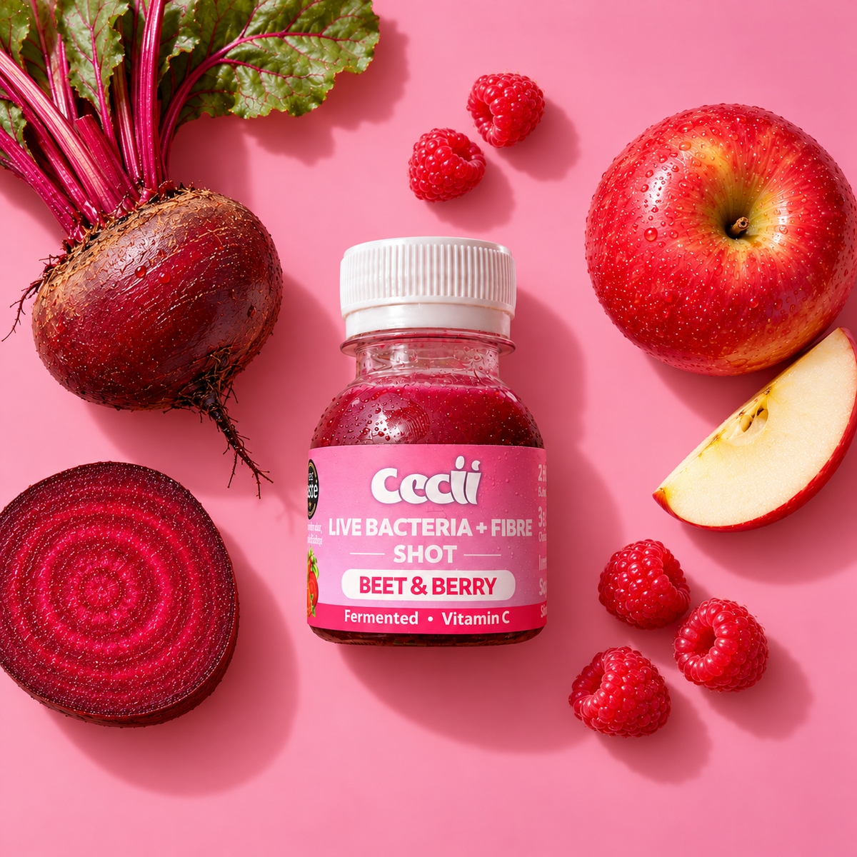 A bottle of Cecii Beet & Berry Live Bacteria + Fibre Gut Health Shot centered on a pink background, surrounded by fresh ingredients including a whole beet with greens, a sliced beet, a whole apple, and several raspberries.